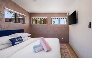 Un dormitorio con papel tapiz estampado, una cama grande con ropa de cama blanca, un cabecero azul, un televisor montado en la pared y una ventana con vista a palmeras.
