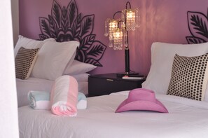 A neatly made bed with pillows, a nightstand with a lamp, and a wall with a leaf design. - The Kate Suite · Two Queens · Palm Springs Pool (Palm Springs)