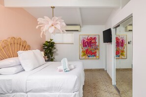 Ein Hotelzimmer mit einem Bett, einer großen, gefiederten Leuchte, einem an der Wand montierten Fernseher und zwei farbenfrohen Gemälden.