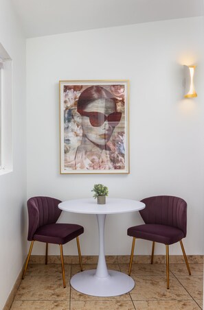 A room with a white round table, two purple chairs, a framed abstract painting, and a wall-mounted light fixture. - Brigitte Suite · Adults-Only PS Boutique· Dog Yard (Palm Springs)