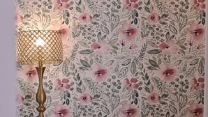 A room with floral wallpaper, a white dresser with drawers, a beige armchair, a small ottoman, and a table lamp.