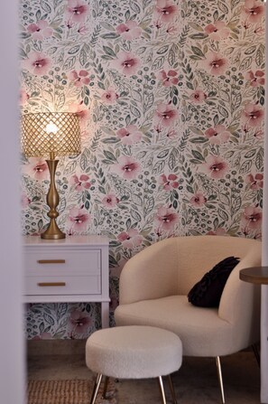 A room with floral wallpaper, a white dresser with drawers, a beige armchair, a small ottoman, and a table lamp. - Brigitte Suite · Dog-Friendly Yard, Palm Springs (Palm Springs)