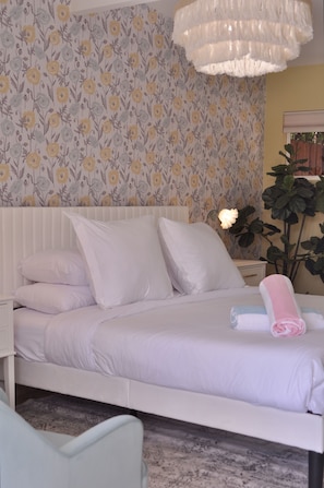 A neatly made bed with white bedding, a bedside table with a lamp, and a floral wallpaper pattern. - The Audrey Suite · Dog-Friendly Yard & Pool, PS (Palm Springs)