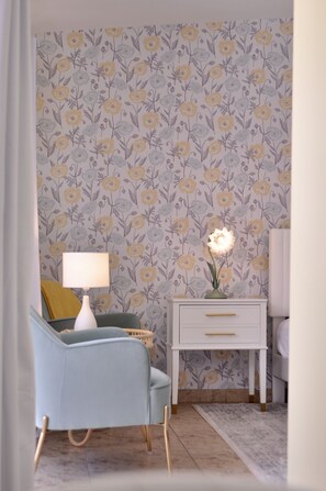 A room with floral wallpaper, a light blue armchair, a bedside table with a lamp, and a nightstand with a vase of flowers.