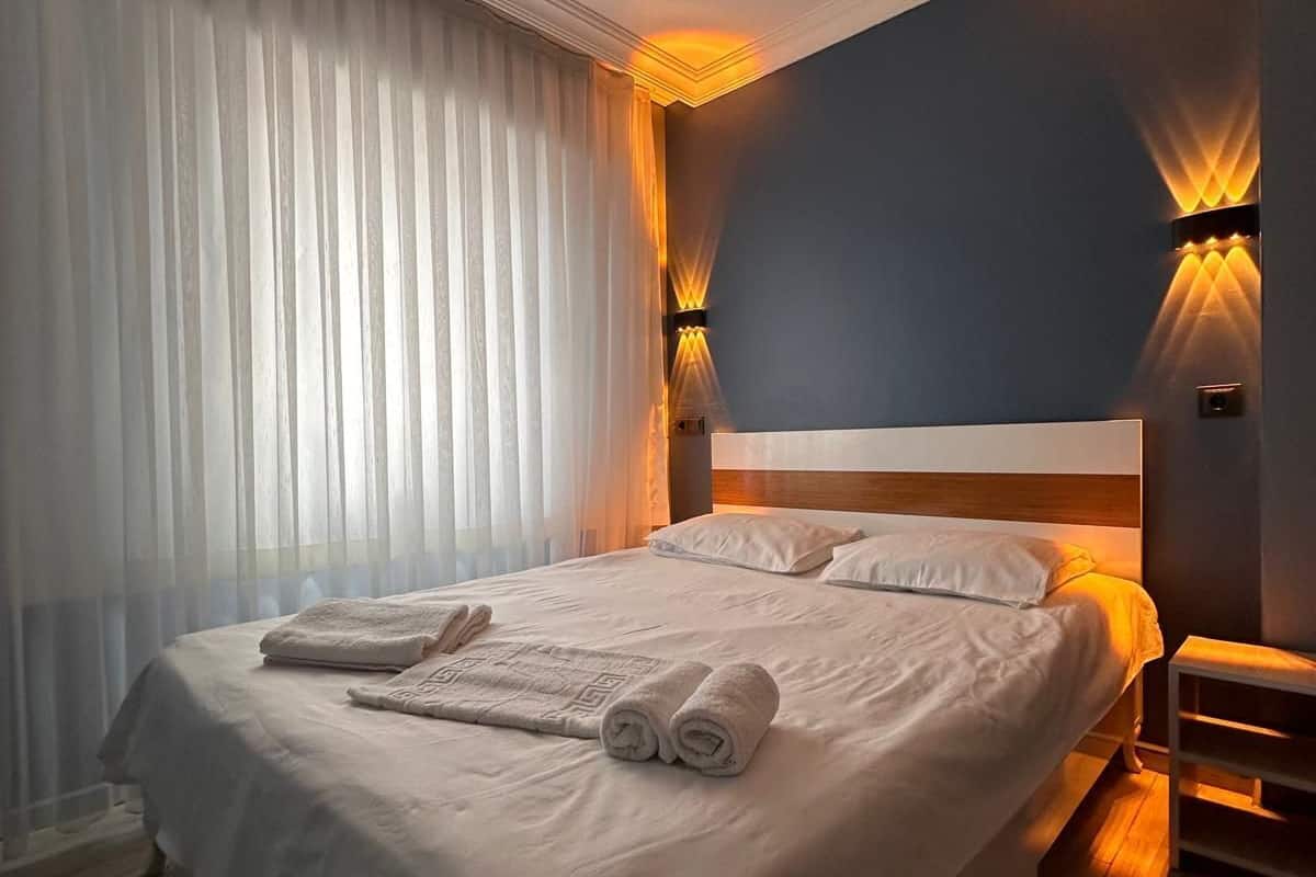 A neatly made bed with white linens and rolled towels, a headboard, and two wall-mounted light fixtures.