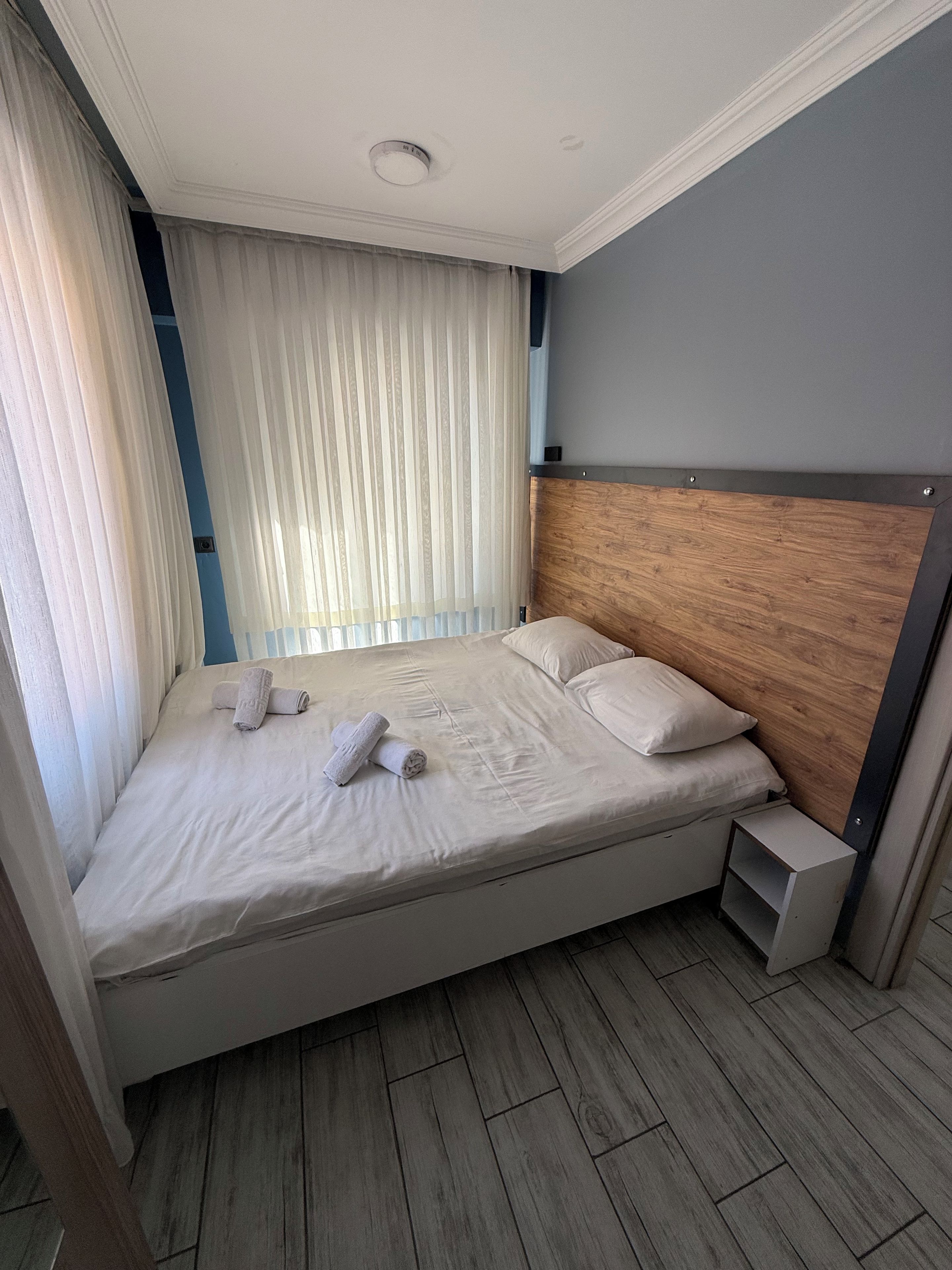 Una cama bien hecha con almohadas y toallas enrolladas, cabecera de madera, mesita de noche y ventana con cortinas traslúcidas.