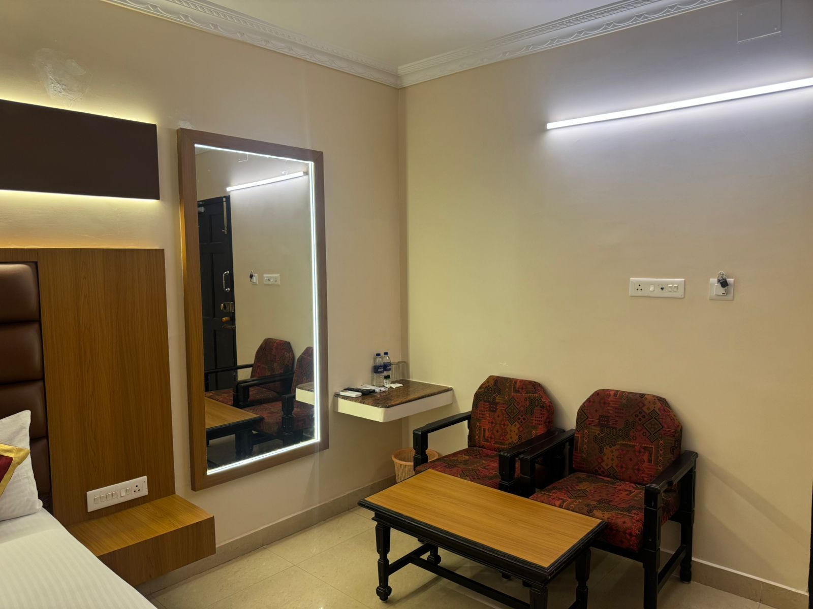 A hotel room with a bed, a bedside table, a mirror, two patterned chairs, a small table, and a nightstand.