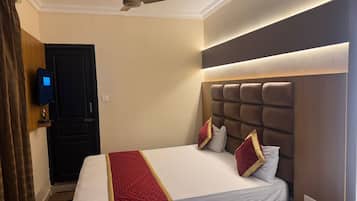 Una habitación de hotel con cama, ventilador de techo, televisor montado en la pared y mesita de noche.