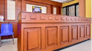 A reception desk with a blue chair, a ceiling fan, and a window with metal bars. - HOTEL SRIMANIYA (Nagercoil)