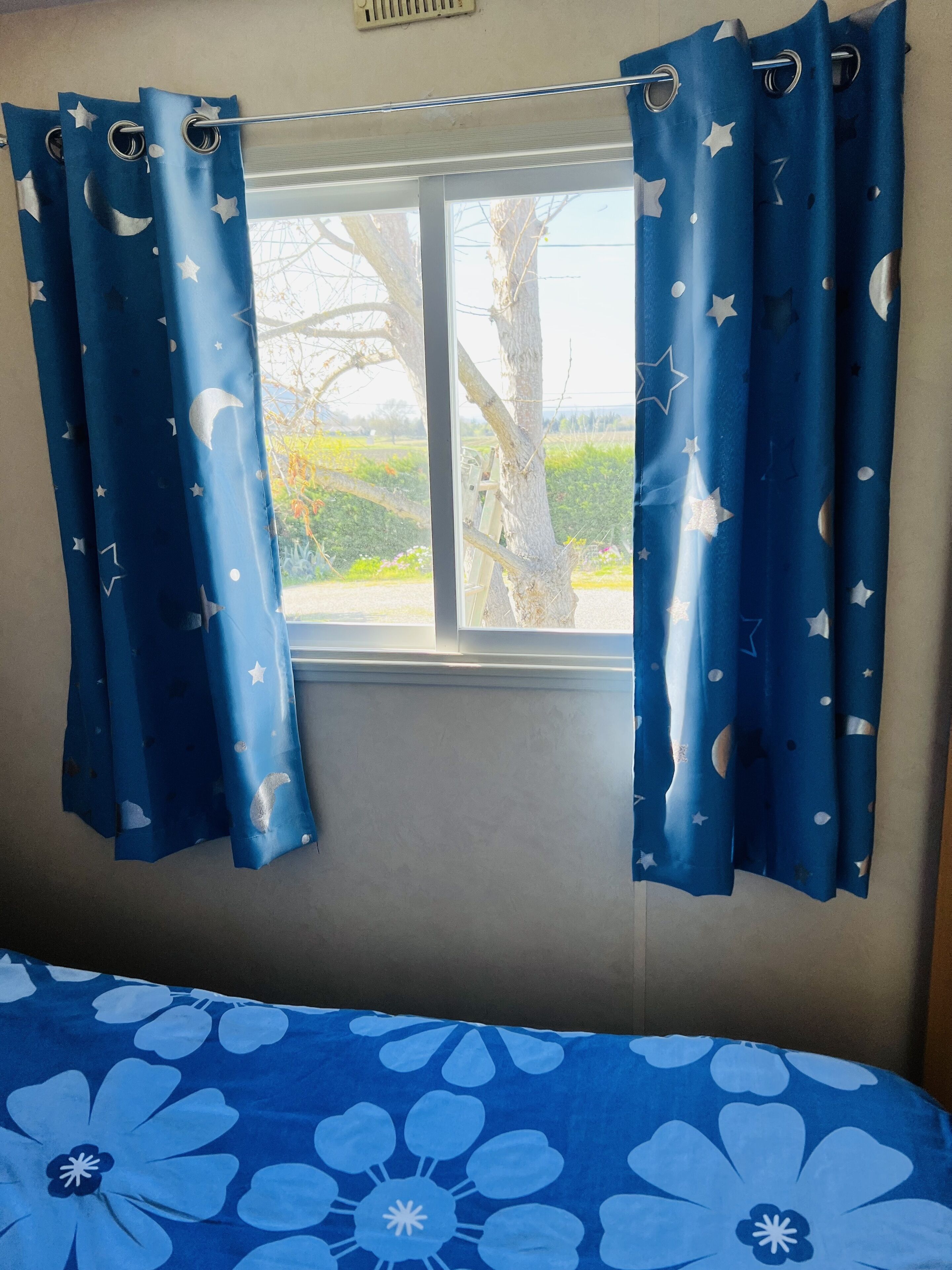 Une pièce avec une fenêtre ornée de rideaux bleus à motifs d’étoiles et de lunes, un lit recouvert d’une couvre-lit à fleurs bleues, et une vue sur des arbres et une route à l’extérieur.
