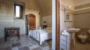 A bedroom with a stone floor, a wooden bed, a nightstand, a small table, and a window. The bathroom includes a sink, a mirror, a bidet, and a towel rack.
