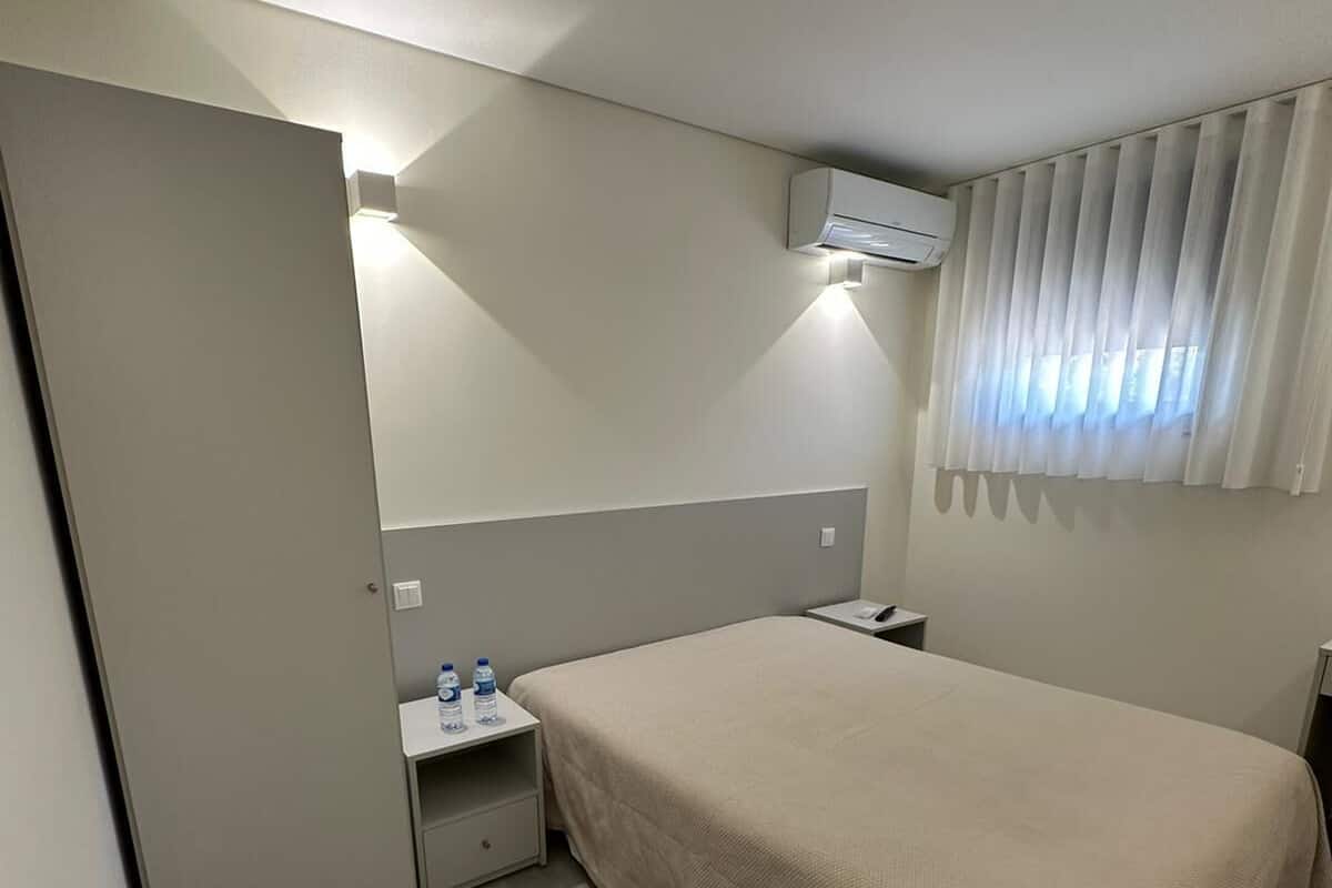 A hotel room with a bed, bedside table, and an air conditioning unit.