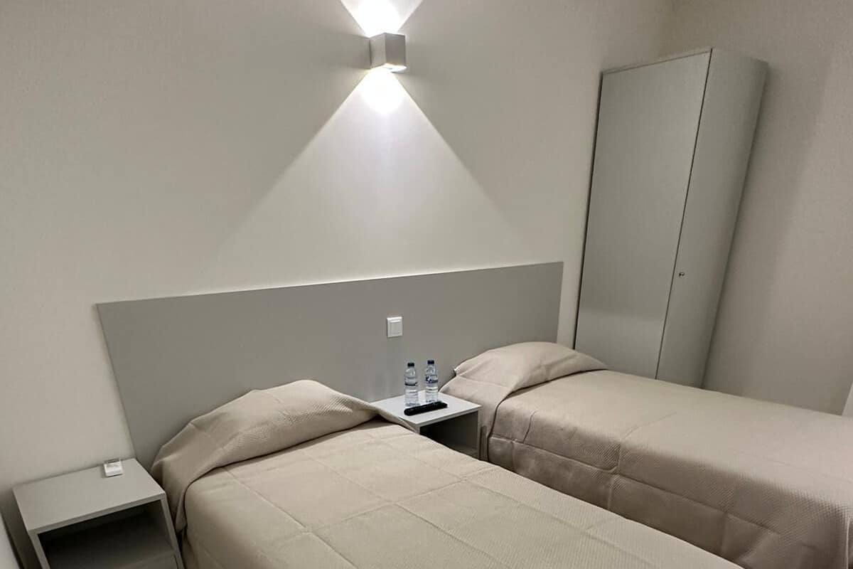 A hotel room with two beds, a wardrobe, a nightstand, and a wall-mounted light fixture.