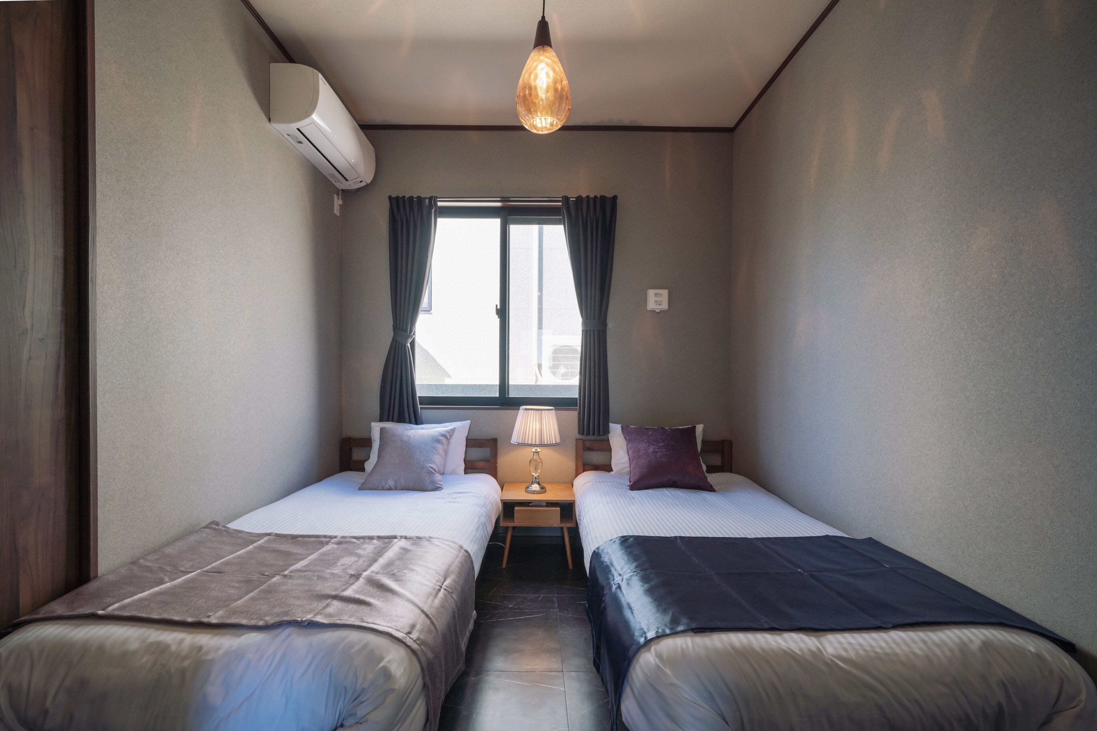 Et lille, moderne værelse med to senge, et vindue med gardiner, et bord med en lampe og en aircondition.