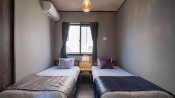Et lille, moderne værelse med to senge, et vindue med gardiner, et bord med en lampe og en aircondition.
