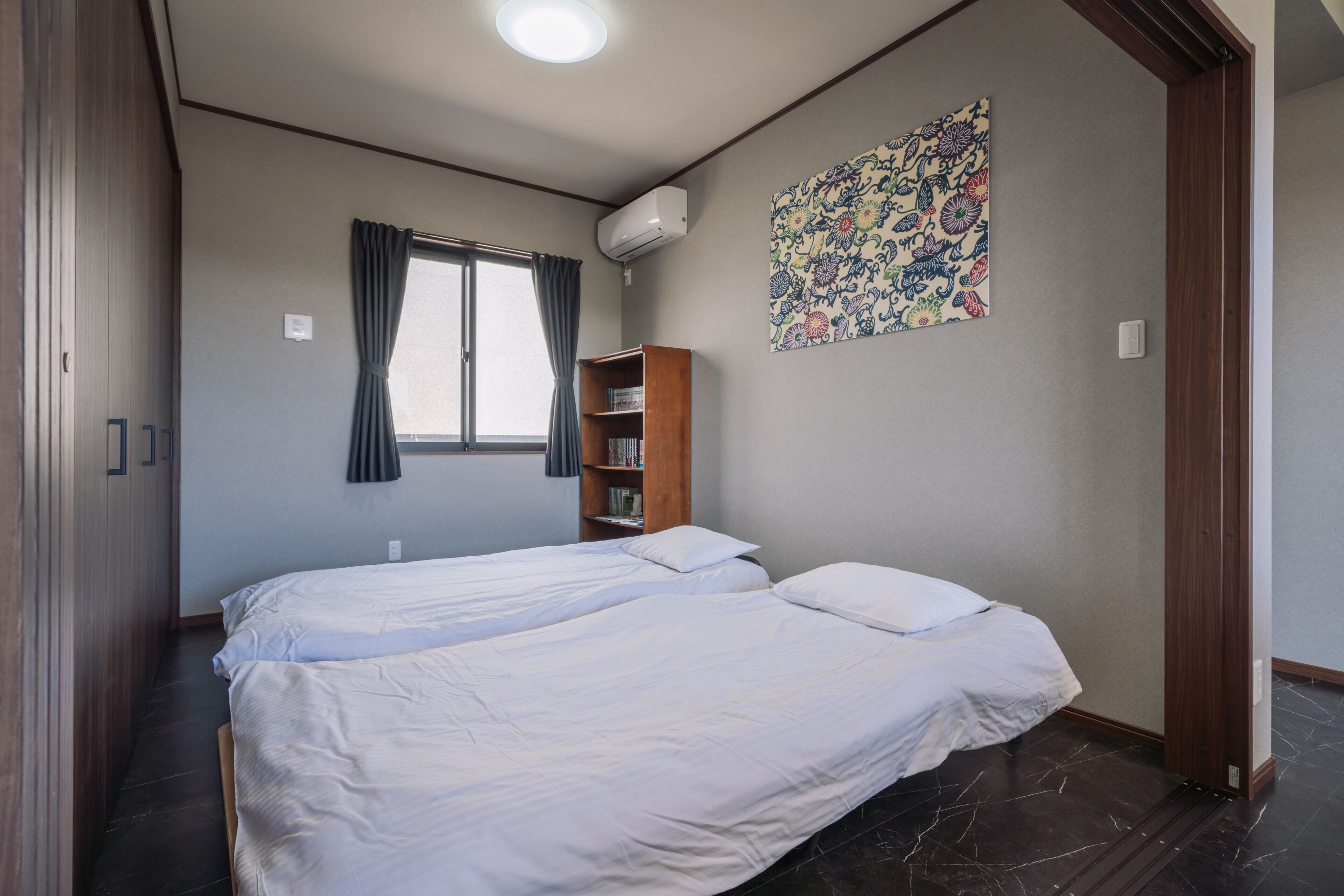 A bedroom with two beds, a window with curtains, a bookshelf, and a floral wall decoration.