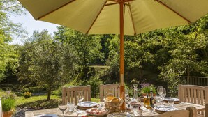 Mesa de comedor al aire libre con platos, vasos y una cesta, rodeada de sillas, bajo un paraguas grande, con un jardín exuberante al fondo.