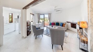 A spacious living area with a sofa, armchairs, and a coffee table. A kitchen is visible in the background, and there is a large window with curtains. - La Manga Club El Rancho Townhouse 513 (Atamaría)