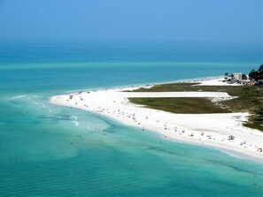 Una playa costera con una amplia zona de arena, aguas turquesas cristalinas y algunos edificios al fondo.