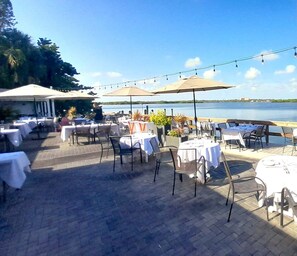 Un área de comedor al aire libre con mesas, sillas, sombrillas y vistas al agua.
