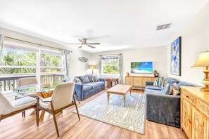 A spacious living room with a ceiling fan, large windows, a sofa, a coffee table, and a view of the outdoors.