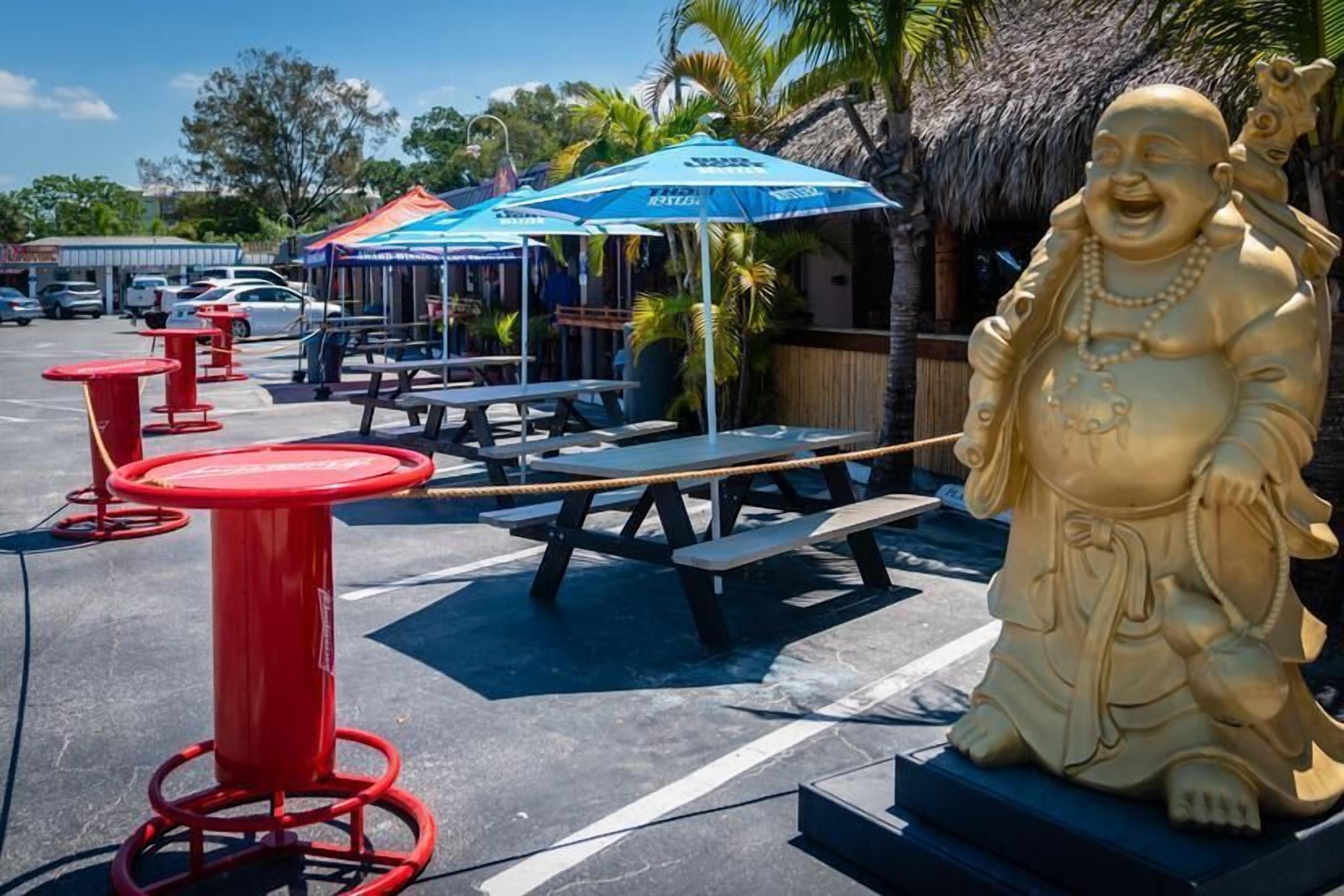 A statue of a laughing Buddha, outdoor seating with tables and umbrellas, and a parking area with cars.