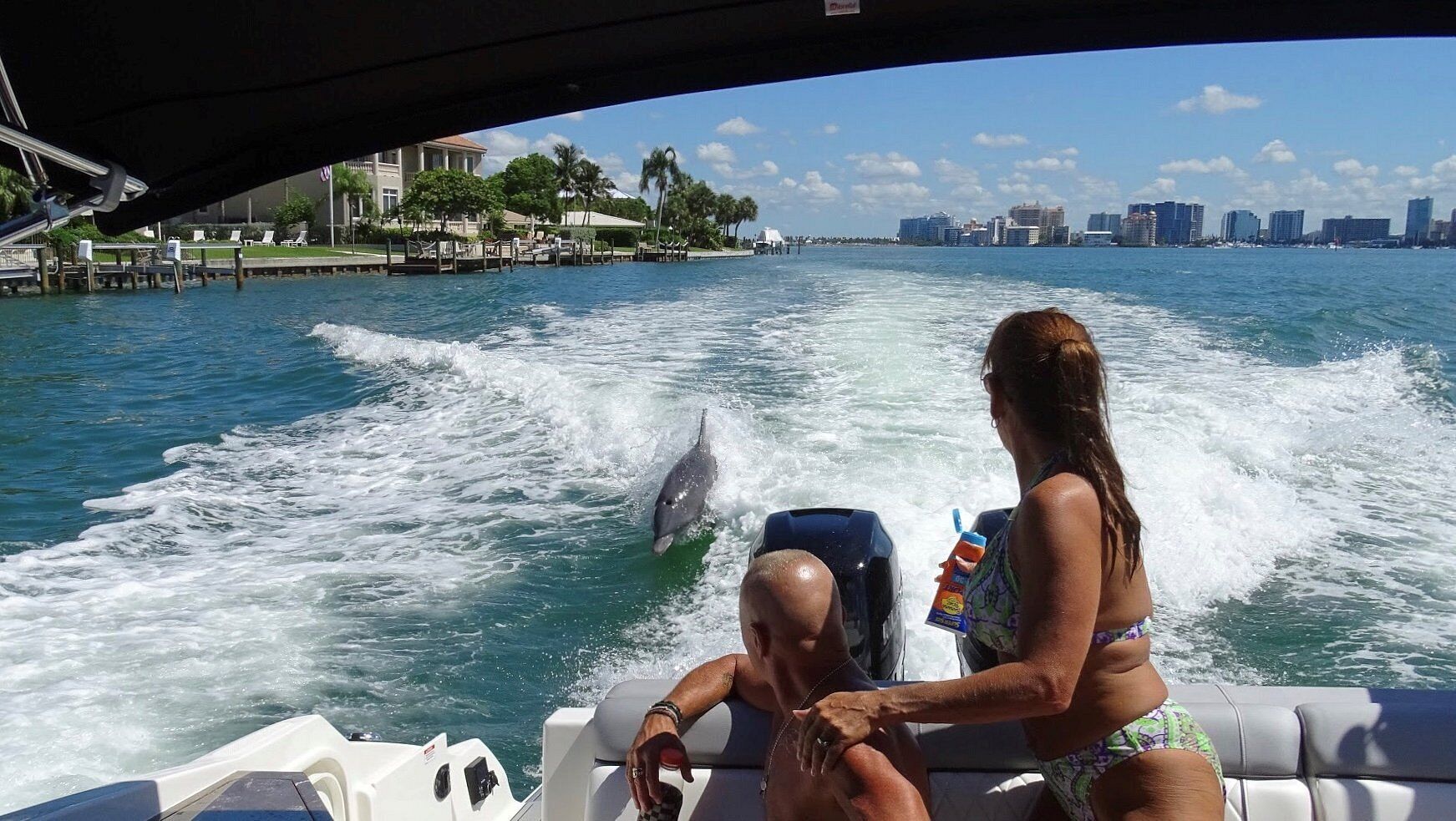 A boat with two people, one holding a bottle, and a dolphin jumping in the water.