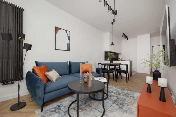 A modern living room with a blue sofa, a round coffee table, and a dining area with a table and chairs. - Elegant Apartment with Balcony | Close to Metro, Place to Work (Warszawa)