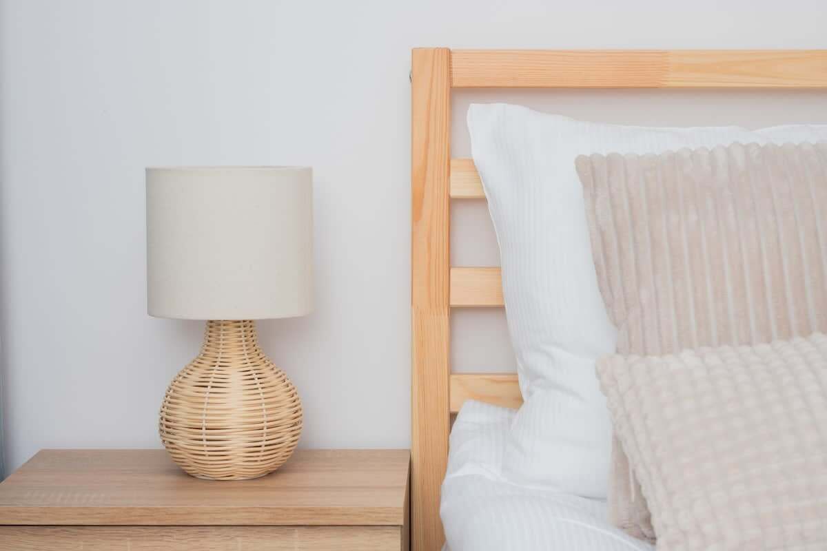 Een net opgemaakt bed met wit en beige beddengoed, een houten hoofdbord en een rieten lamp op een houten nachtkastje.