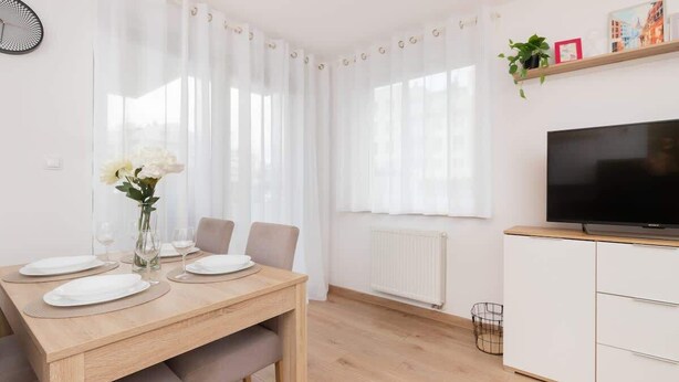 A modern living room with a dining table set for four, a flat-screen TV, and a radiator. - Apartment with Garden | Place to Work | Close to the Lake (Gdansk)