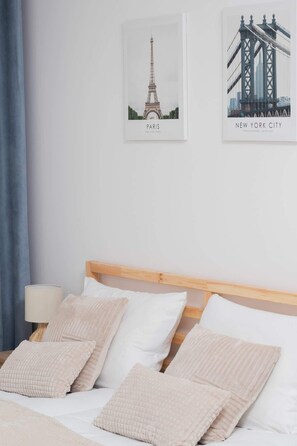 A neatly made bed with white linens and beige pillows, a wooden headboard, and two framed pictures of the Eiffel Tower and the Brooklyn Bridge.