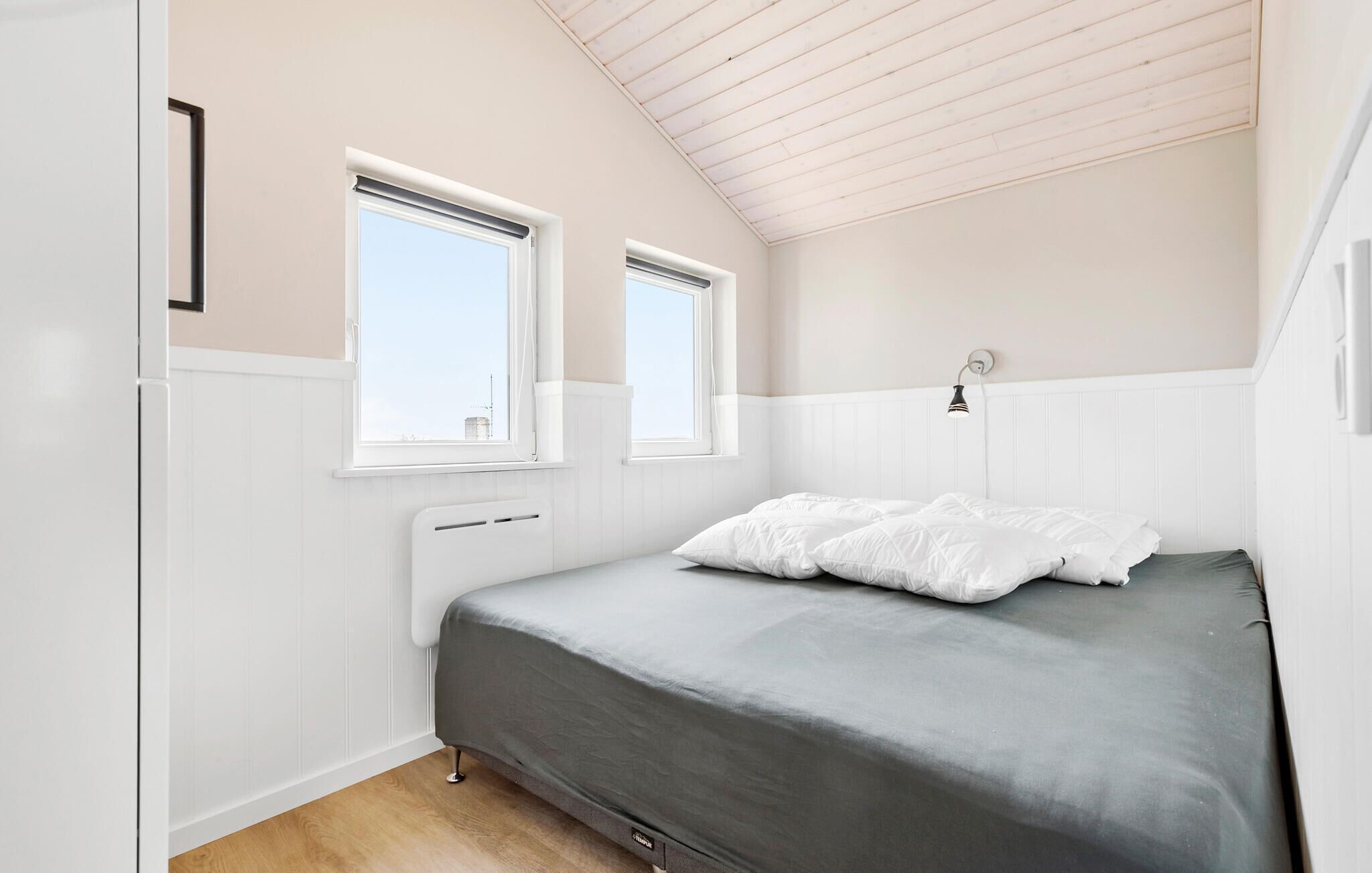 Et soveværelse med skråvæg, to vinduer, en seng med hvid og grå sengetøj, en sengetavlelampe og en radiator.