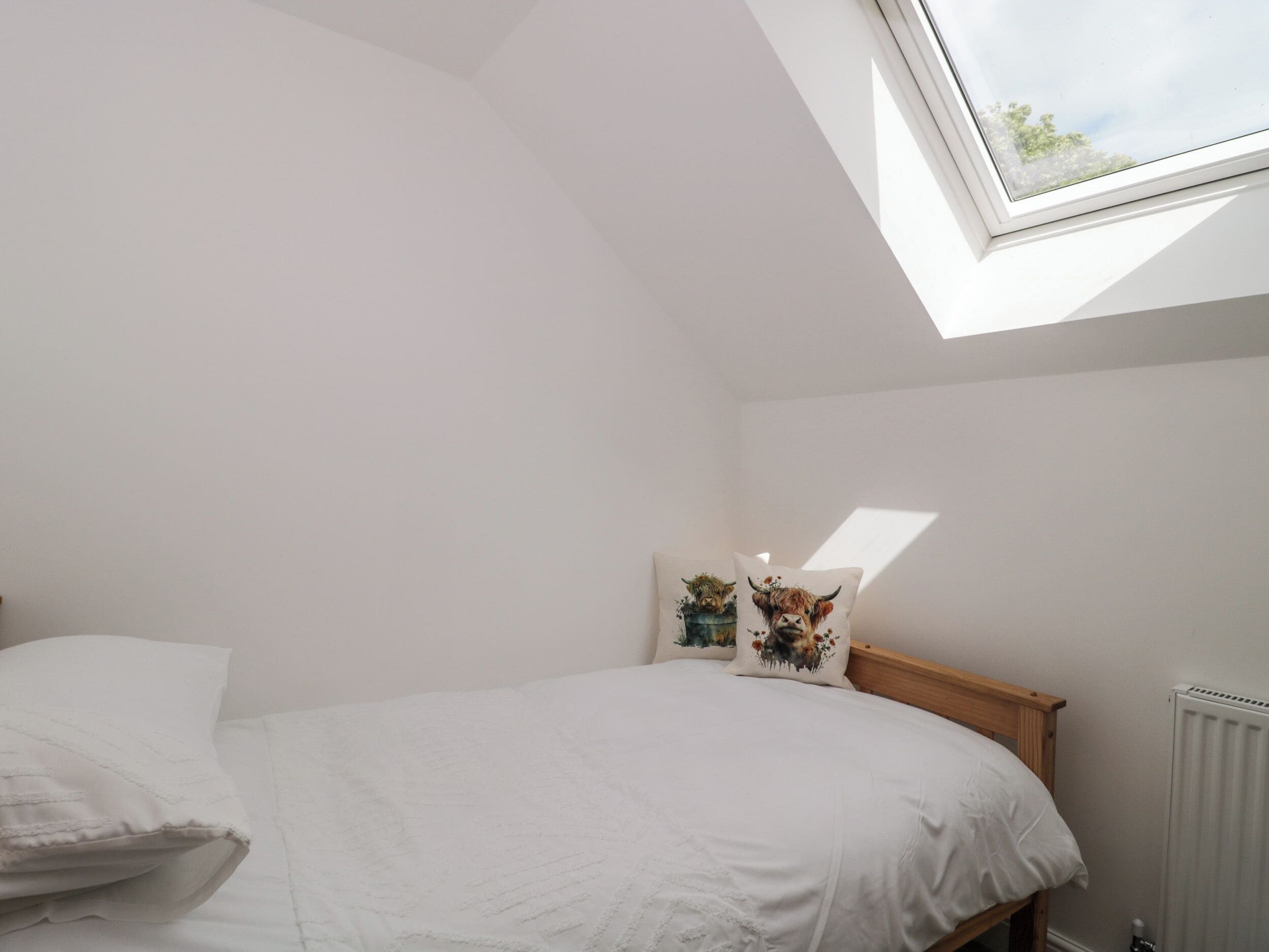 Quarto com claraboia, cama com roupa de cama branca e dois travesseiros decorativos com estampas florais.