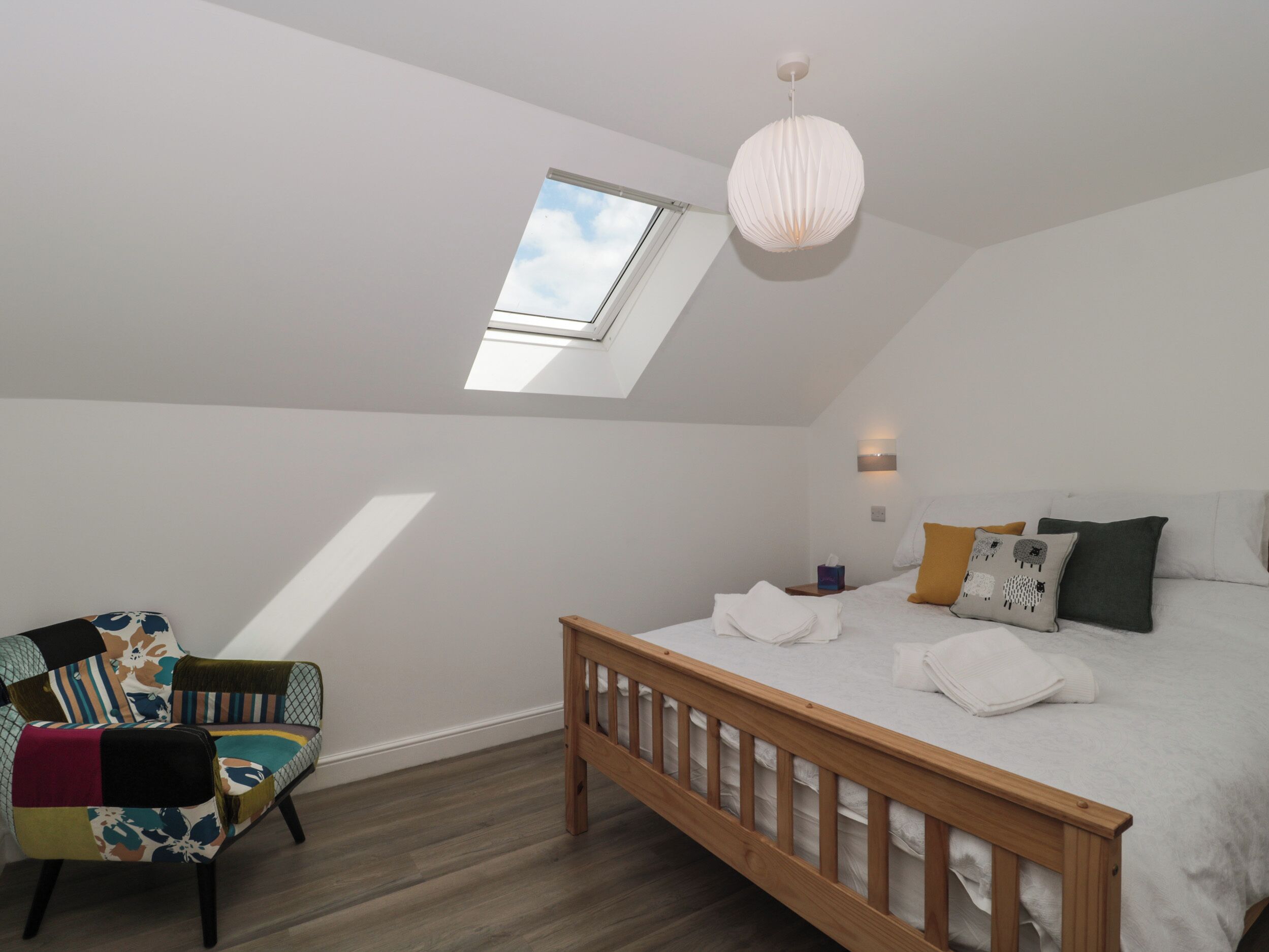 A bedroom with a skylight, a bed with white linens, a colorful chair, and a wooden headboard.