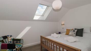 A bedroom with a skylight, a bed with white linens, a colorful chair, and a wooden headboard.