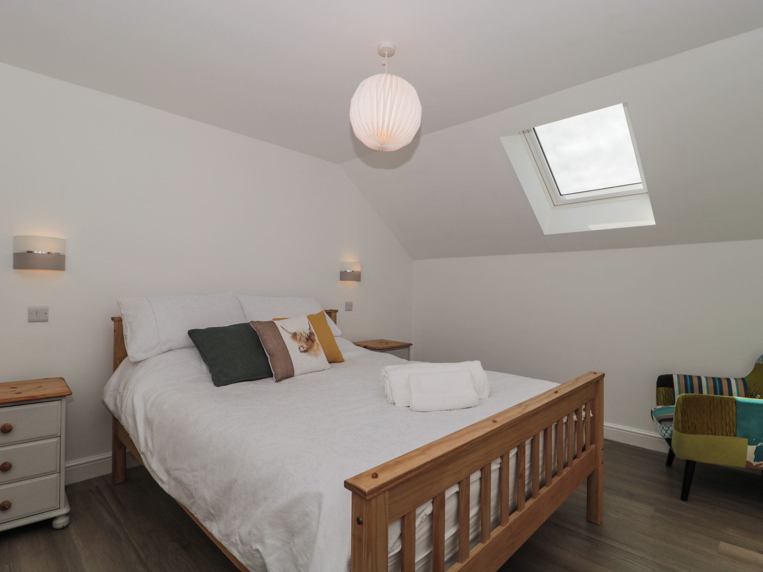 A bedroom with a wooden bed, white bedding, a skylight, and a small bedside table with a lamp.