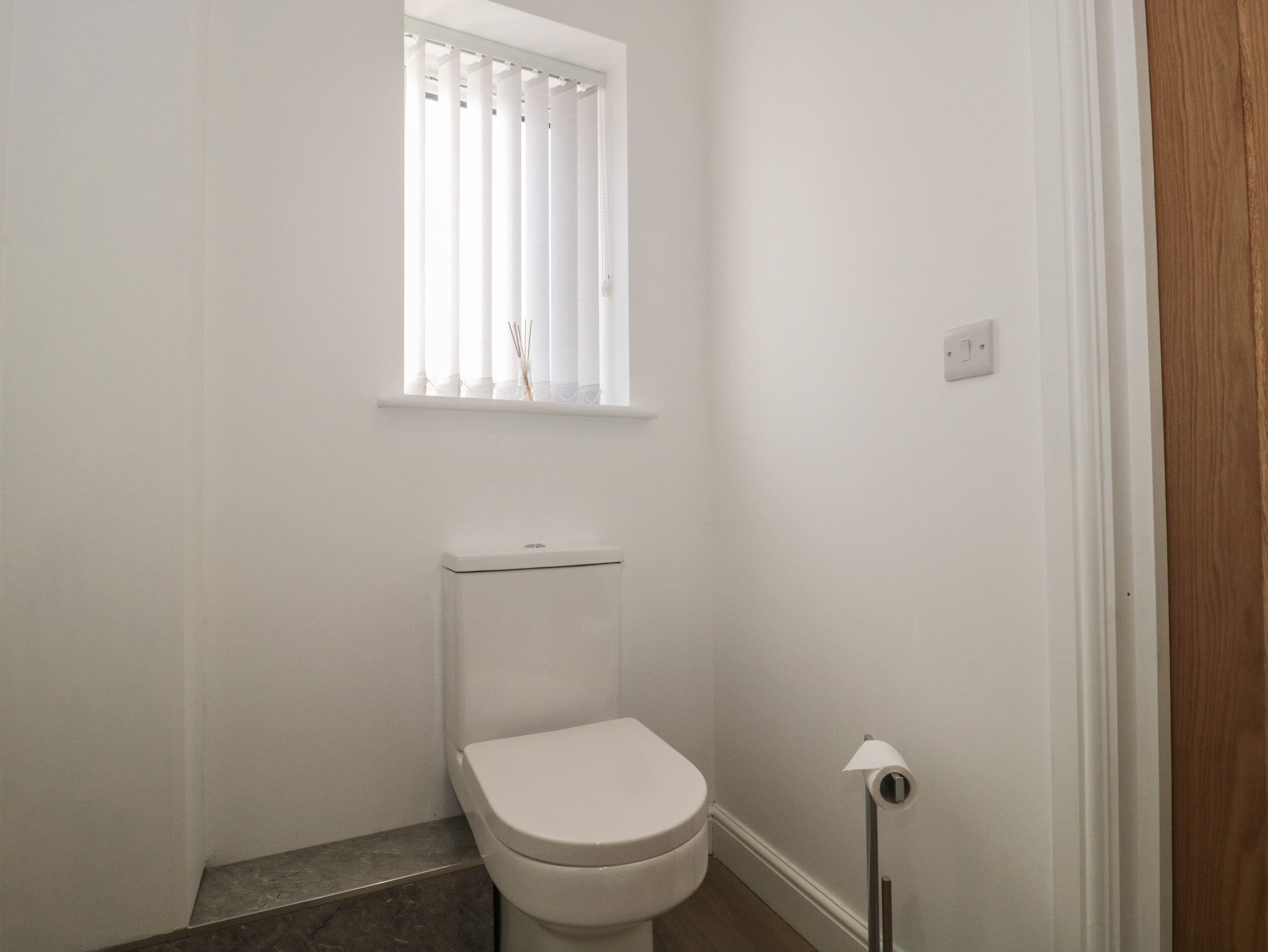 A small, white bathroom with a toilet, a window with blinds, and a toilet paper holder.