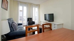 En kompakt opholdsstue med sofa, spisebord, fjernsyn og en balkon med gardiner.