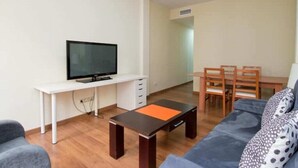 En moderne stue med et fladskærms-TV på et hvidt skrivebord, et træspisebord med stole og en grå sofa med mønstrede puder.