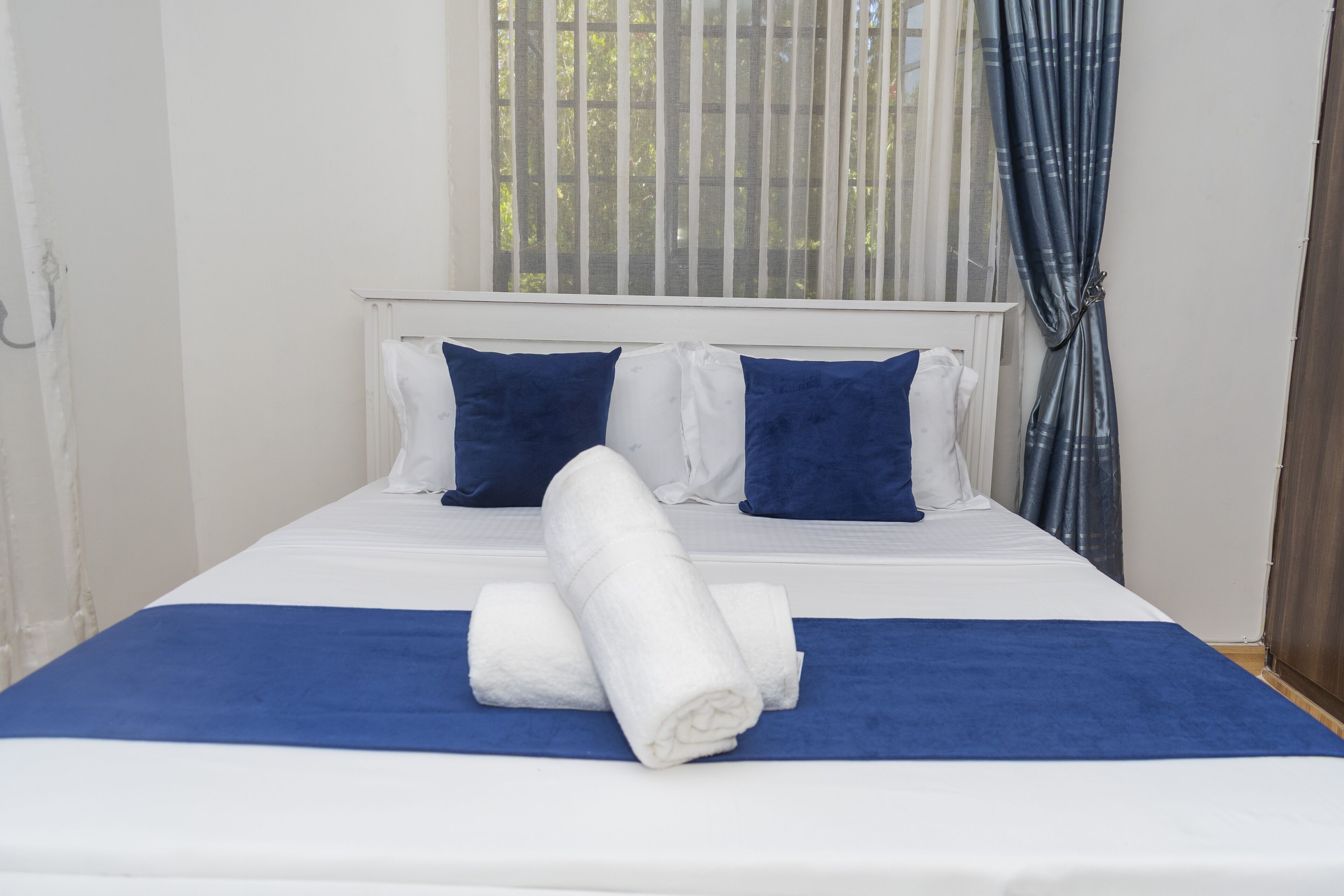 Een net opgemaakt bed met wit en blauw beddengoed, twee kussens en opgerolde handdoeken.
