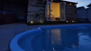 A modern house with a swimming pool, wooden exterior, and a well-lit interior visible through the open door. - Private Pool, Hot Tub, Luxury Cabana Poole Dorset. (Poole)