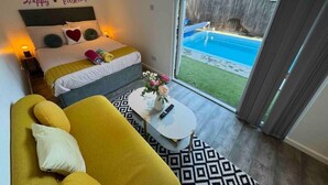 A bedroom with a bed, a yellow sofa, a small table with a vase, and a view of a backyard swimming pool. - Private Pool, Hot Tub, Luxury Cabana Poole Dorset. (Poole)