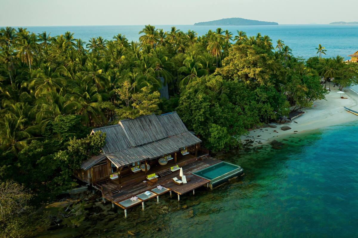 An aerial view of a wooden house with a thatched roof, surrounded by dense palm trees, near a clear blue sea with a sandy beach and a small boat.