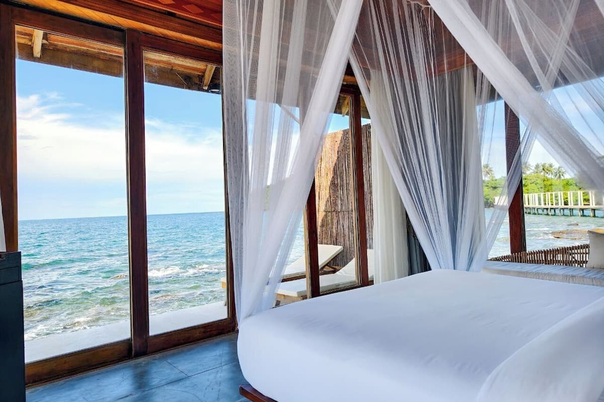 A bedroom with a large bed, white bedding, and a view of the ocean through sheer curtains.