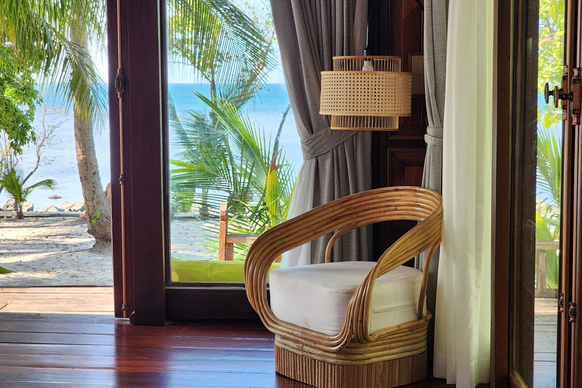 Uma cadeira de vime com almofada em um quarto com piso de madeira, vista para a praia e oceano através de uma janela, e uma luminária de rattan.