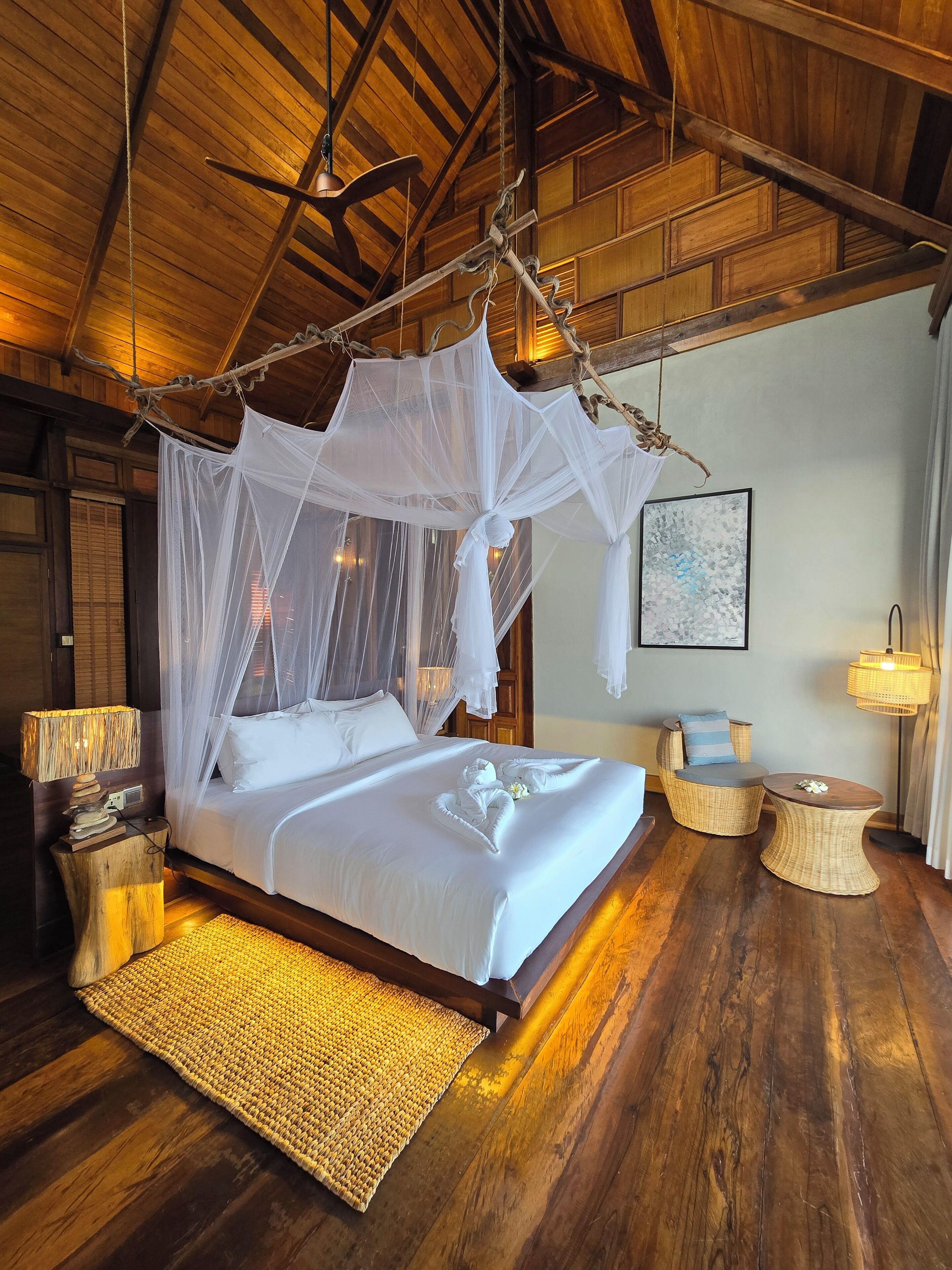 A spacious bedroom with a large bed, a canopy bed, wooden flooring, a woven rug, a bedside table with a lamp, and a chair.