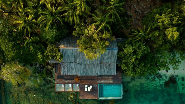 An aerial view of a wooden overwater bungalow with a swimming pool, surrounded by lush greenery and clear turquoise waters. - Full Moon Private Island (Kiri Sakor)