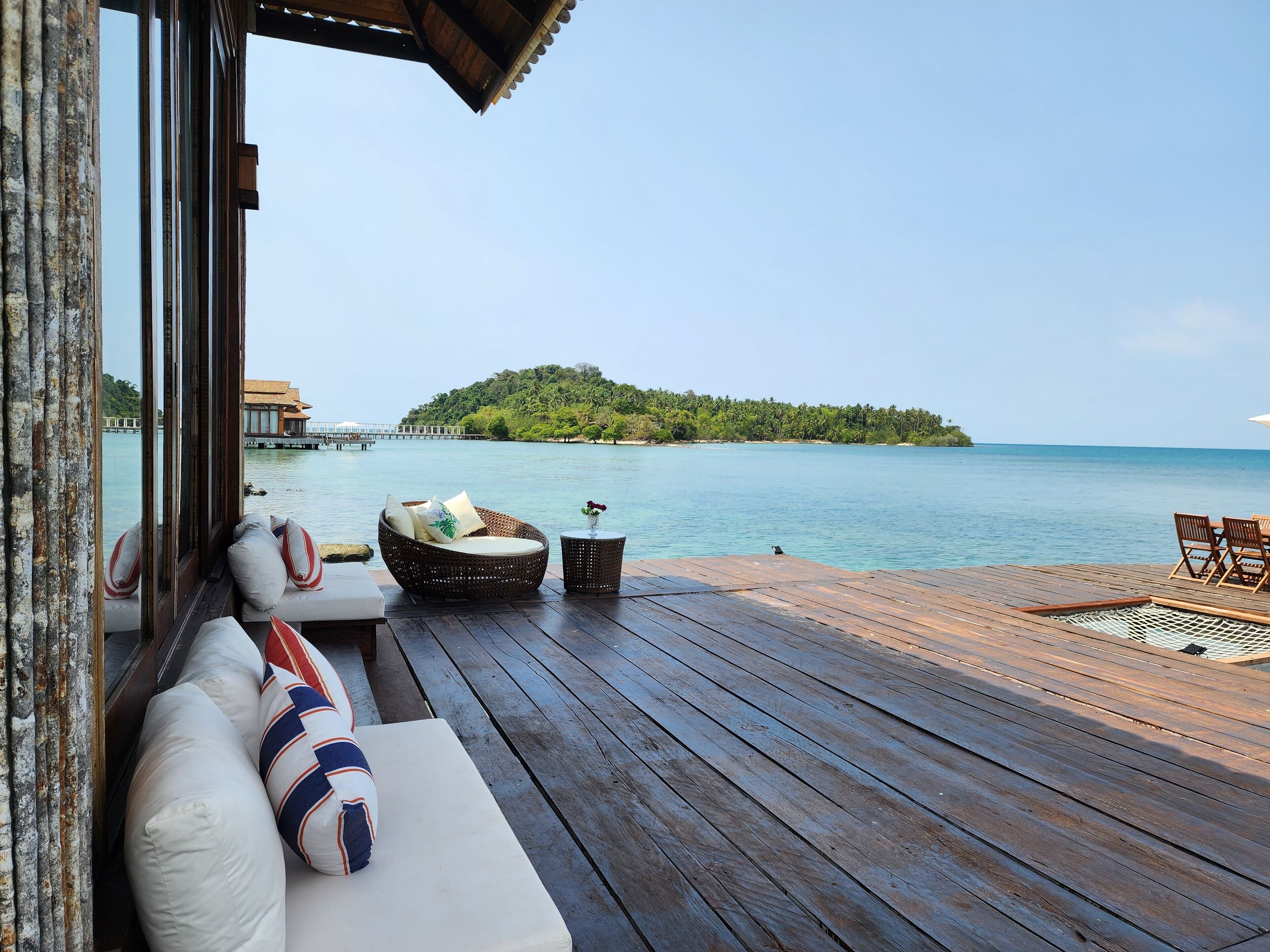 A wooden deck with a wicker sofa and a small table, overlooking a calm sea and a lush island.