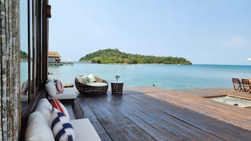 A wooden deck with a wicker sofa and a small table, overlooking a calm sea and a lush island.