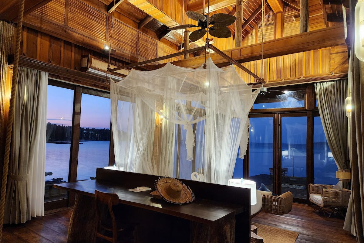 A room with a wooden ceiling, a large window overlooking a body of water, a table with a hat, and a canopy.